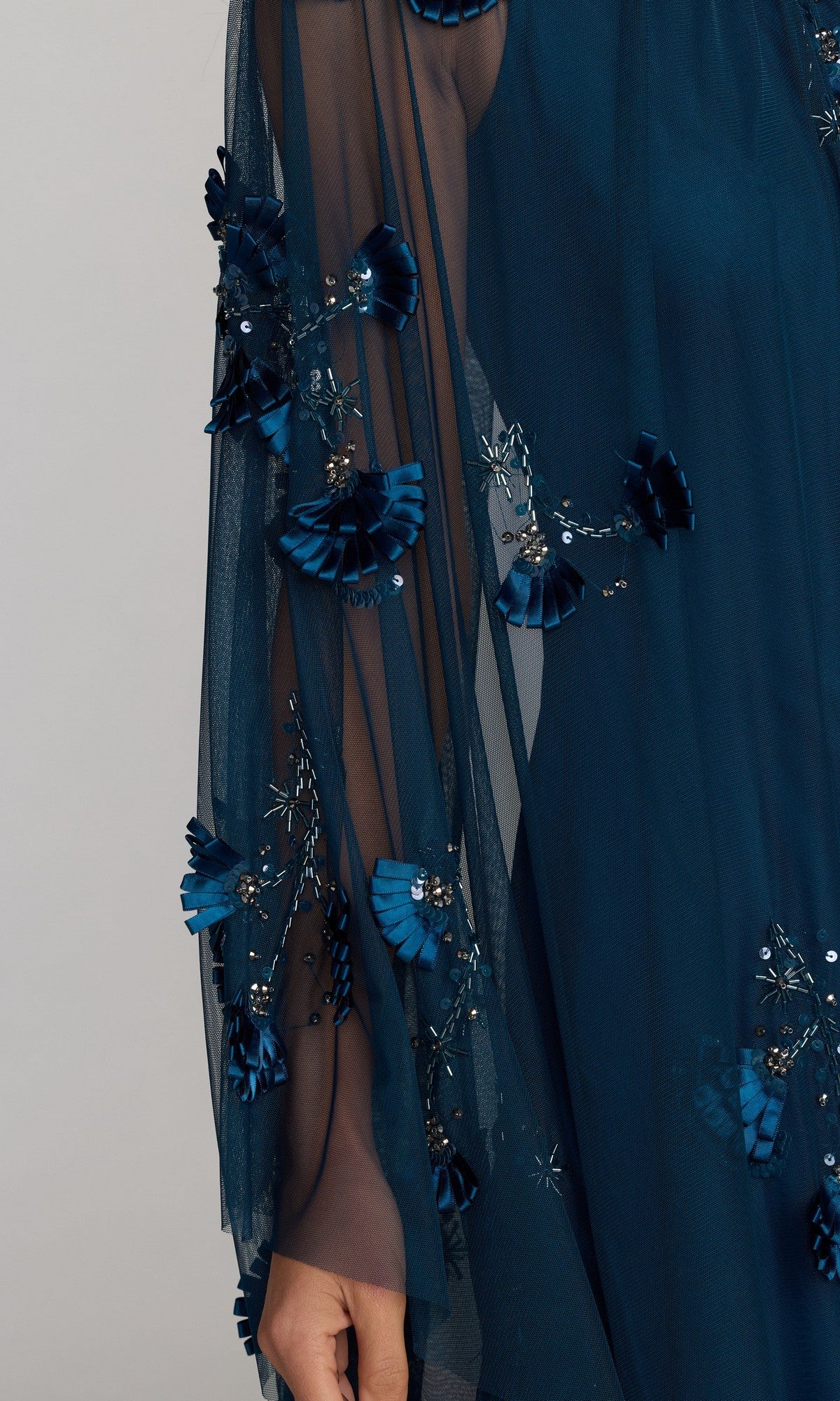 Close-up of sheer dark blue fabric adorned with 3D floral appliqués, sequins, and beads, with a person's hand partially visible beneath—an elegant detail from the Long Formal Dress: Mac Duggal 9374.
